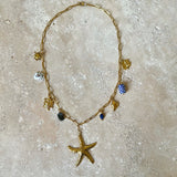 Gold necklace with starfish pendant, blue Apatite and Labradorite Gemstone, sun charm, lucky charm, turtle, shell and delft blue ceramic worn by a person in a white shirt