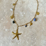Gold necklace with starfish pendant, blue Apatite and Labradorite Gemstone, sun charm, lucky charm, turtle, shell and delft blue ceramic worn by a person in a white shirt