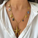 Gold necklace with starfish pendant, blue Apatite and Labradorite Gemstone, sun charm, lucky charm, turtle, shell and delft blue ceramic worn by a person in a white shirt